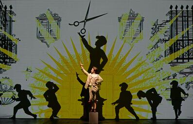 Vergrößerte Ansicht von Ilya Lapich (Figaro) steht heroisch auf einem Podest, über ihm ist eine Schere an die Wand projiziert.