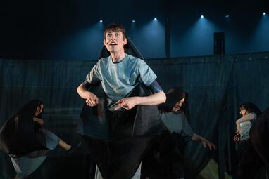 Vergrößerte Ansicht von Ein Mann in einem schwarzen Umhang im Vordergrund. Drei Menschen tanzen hinter ihm.