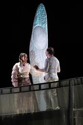 Vergrößerte Ansicht von Frau in rosa redet mit Mann in weiß vor einem großen Libellenflügel.