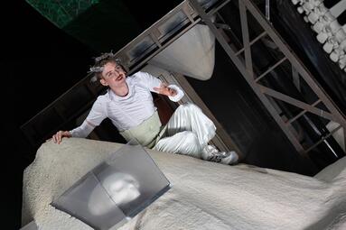 Vergrößerte Ansicht von Person in weiß, mit Lockenwicklern im Haar, sitzt auf einem Sandberg. Auf dem Sand liegt ein Totenschädel in einer Plastikkiste.