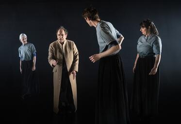 Vergrößerte Ansicht von Drei Personen in blauen Shirts und schwarzen Hosen schauen einen Mann in einem braunen Mantel verwirrt an.