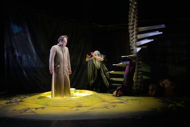 Vergrößerte Ansicht von Dramatisches, gelbes Licht fällt auf einen Mann in einem braunen Mantel. Person mit langen, grauen Haaren steht neben einer Wendeltreppe, die wie eine Wirbelsäule aussieht