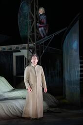 Vergrößerte Ansicht von Mann in langem, braunen Mantel steht im Vordergrund. Person in schwarz-rot hängt an einem metallenen Gestell im Hintergrund.