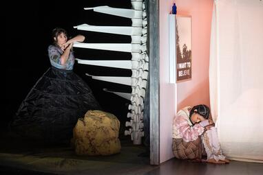 Vergrößerte Ansicht von Frau lehnt sich an eine Wendeltreppe, die wie eine Wirbelsäule aussieht. Sie wird von einer rosa Wand von einer anderen Frau getrennt. Diese sitzt trauernd unter einem Bild mit den Buchstaben &quot;I WANT TO BELIEVE&quot;.