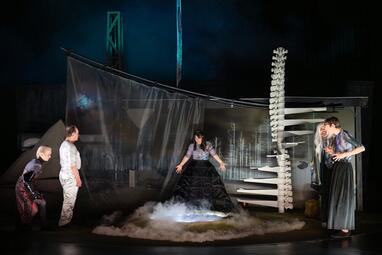 Vergrößerte Ansicht von Fünf Personen stehen um ein dampfendes Loch im Boden. Hinter ihnen hängt eine schwarz-weiße Baumlandschaft, ein transparenter Vorhang und eine Wendeltreppe, die wie eine Wirbelsäule aussieht.