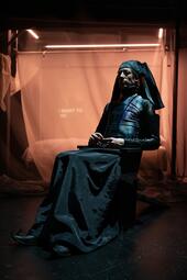 Vergrößerte Ansicht von Dramatisches orange-blaues Licht fällt auf eine Person in einem schwarzen Rock und schwarzer Haube.