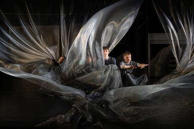 Vergrößerte Ansicht von Eine Gruppe Menschen sitzen auf einem flatternden, transparenten Tuch, das sie umhüllt.