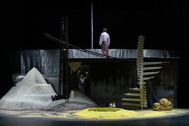 Vergrößerte Ansicht von Bühnenbild zeigt zwei Ebenen, die mit einer Wendeltreppe, die wie eine Wirbelsäule aussieht, verbunden wird. Auf der oberen Ebene steht eine Person, die der Kamera den Rücken zudreht. Neben der Plattform befindet sich ein Sandhaufen.