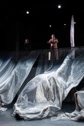 Vergrößerte Ansicht von Zwei Personen schauen von einer erhöhten Plattform auf lange, flatternde, glänzende Tücher.