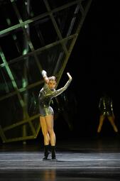 Vergrößerte Ansicht von Eine Person in einem silbernen Jumpsuit und schwarzen Socken steht auf den Zehenspitzen und hält eine Hand hoch an ihren Kopf und streckt die andere vor sich in die Luft, sodass sie ihre Handflächen anschaut.