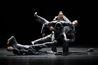 Vergrößerte Ansicht von Es sind vier Personen zu sehen in grauen Anzügen. Die erste Person liegt seitlich auf dem Boden und streckt einen Arm und ein Bein in die Luft. Links neben ihm steht der zweite Mann mit gebeugten Knien und schaut auf den Boden. Hinter ihm steht ein…