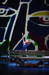 Vergrößerte Ansicht von Im Vordergrund liegt ein Mann mit einer dunklen Hose bekleidet bäuchlings unter einer weißen Bank und hebt Arme und Beine in die Luft. Hinter der Bank steht eine Frau in einem bunten Kleid und hebt die Arme hoch. Sie schaut zur Decke. Es sind…