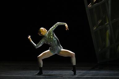 Vergrößerte Ansicht von Eine Person steht breitbeinig in einem silbernen Jumpsuit auf der Bühne und hebt die Arme angewinkelt von sich.