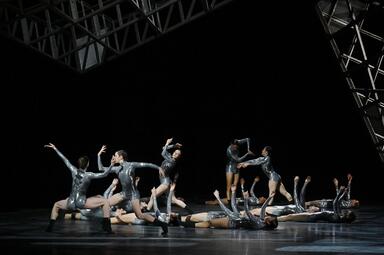 Vergrößerte Ansicht von Es sind etwa fünfzehn Personen in glänzenden grauen Kostümen zu sehen. Manche tanzen, während andere auf dem Boden liegen und die Arme in die Luft strecken.