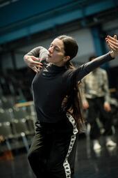 Vergrößerte Ansicht von Es ist eine Person mit braunen langen Haaren in einem schwarze Oberteil und einer schwarzen Hose zu sehen, die leicht in die Knie geht und enen  Arm vollkommen zur Seite ausgestreckt hat. Den anderen Arm winkelt sie zur Häftte an.