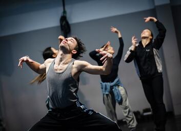 Vergrößerte Ansicht von Im Vordergrund ist eine Person zu sehen, die breitbeinig steht und mit geschlossenen Augen die Arme von sich abwinkelt. Im Hintergrund sind drei weitere Tänzer zu sehen in dunkler Kleidung, die alle in unterschiedliche Richtungen schauen und die…