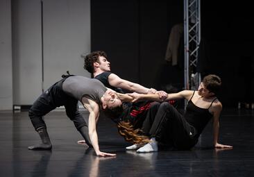 Vergrößerte Ansicht von Auf der Bühne sind drei Personen zu sehen. Eine Person formt mit ihrem Körper eine Brücke, wobei sie sich nur mit einem Arm auf den Boden stützt und sich mit anderen Armen an den anderen Personen festhält. Die andere Person sitzt auf dem Boden und…