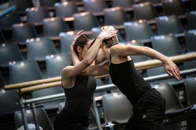 Vergrößerte Ansicht von Zwei Personen halten ihre Gescihter nah aneinander und halten sich mit den Händen am Kopf der anderen Person fest. Dabei berühren sich ihre Oberkörper nicht.Beide haben schwarze Kleidung an.