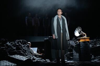 Vergrößerte Ansicht von Ein Mann steht alleine in den Ruinen neben einem Grammophon. Er trägt zeitlose Kleidung, einen dunklen Mantel und hellgrauen Schal über den Schultern. Die Stimmung ist dystopisch, was durch den Nebel verstärkt wird.