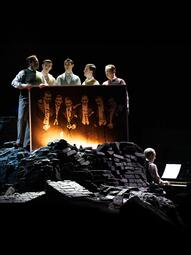 Vergrößerte Ansicht von Auf einem schwarzen Backsteinhaufen stehen fünf Männer unt halten gemeinsam ein signiertes Bild von sechs anderen Männern vor sich. Sie wirken melancholisch und schauen sich gegenseitig an.