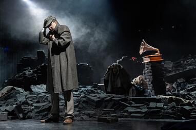 Vergrößerte Ansicht von Ein einzelner, vermummter Mann steht mit dem Rücken zu einem Grammpohon, vor dem zwei andere Personen sitzen. Der stehende Mann hat einen langen, grauen Mantel an und zieht sich die graue Batschkapp tiefer in die Strin, während die anderen Schals…