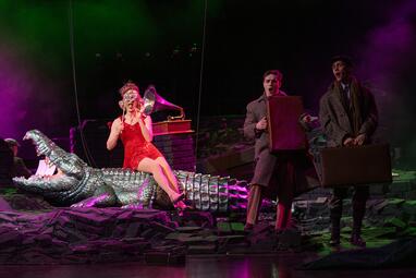 Vergrößerte Ansicht von Eine Frau in einem kurzen, schillernden, roten Trägerrkleid hält sich eine Halbmaske vor das Gesicht. Sie sitzt auf einem Alligator, welcher das Maul aufreißt. Hinter hr steht ein Grammophon auf einer Säule aus schwarzen Backsteinen, neben dem…