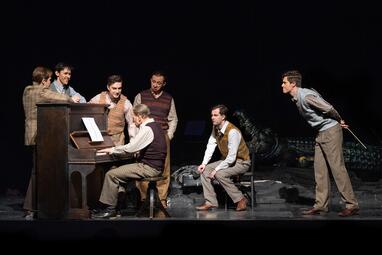 Vergrößerte Ansicht von Ein älterer Mann sitzt links im Bild an einem Klavier und spielt konzentiert. Um ihn herum stehen vier jüngere Männer und lauschen ihm fröhlich. Ein weiterer sitzt gespannt auf der Stuhlkante, auf einem Stuhl schräg hinter dem Pianisten. Er wirkt…