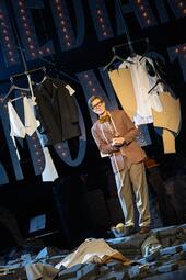 Vergrößerte Ansicht von Ein Mann steht alleine auf Mauerruinen. Er ist umgeben von Kleidungsstücken, die zu Anzügen gehören. Auch auf dem Boden liegen Kleidungsstücke. Der Mann selbst trägt eine große Brille mit schwarzer Umrandung, einen braunen Anzug und braune Fliege.…