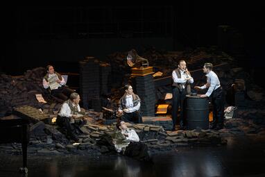 Vergrößerte Ansicht von In den Ruinen von Gebäuden aus schwarzem Stein sitzen vier Männer. Drei von ihnen lesen Zeitung, der vierte sitzt vor einer Schreibmaschine. Zwei weitere Männer stehen an einem großen Fass, einer von ihnen telefoniert mit einem altmodischen…