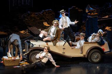 Vergrößerte Ansicht von Vier Männer in weißen Hemden, mit Sonnenbrillen und Strohhüten auf den Köpfen, sitzen in verschiedenen entspannten Posen auf einem beigen Auto. Ein weiterer Mann sitzt neben dem Auto genauso entspannt auf dem Boden. Vor dem Auto wühlt ein anderer…