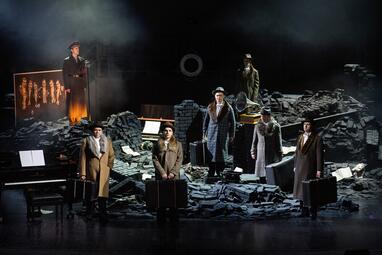 Vergrößerte Ansicht von In den schwarzen Ruinen aus Backsteinen stehen fünf Männer in Reisekleidung, mit Hüten und langen Mänteln. Alle tragen jeweils einen großen Koffer. Im Hintegrrund steht eine weitere Person und beobachtet die Szene. Links steht auf einem Haufen…