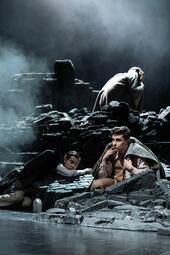 Vergrößerte Ansicht von Im Vordergrund sitzt ein Mann mit einer Decke um die Schulter in den schwarzen Ruinen einer zerstörten Stadt. Links neben ihm wird er von einem anderen Mann halbliegend beobachtet, der einen Mantel über den Schultern hat. Im Hintergrund sitzt eine…