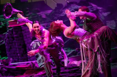 Vergrößerte Ansicht von Drei Schauspieler:innen stehen in expressiven Posen auf einer Theaterbühne mit lila-pinkem Licht. Sie tragen auffällige Kostüme mit Fransen und Glitzerelementen. Im Hintergrund sind aufgetürmte, künstliche Ziegelsteine zu sehen. Links ist ein…