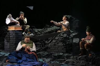 Vergrößerte Ansicht von Vier Schauspieler sitzen und stehen auf einer Bühne, die wie eine Ruinenlandschaft aus schwarzen Ziegelsteinen gestaltet ist. Im Vordergrund sitzt eine Person mit gestrickter Weste und einer Strickarbeit in den Händen, zwei Blechdosen auf dem Kopf…