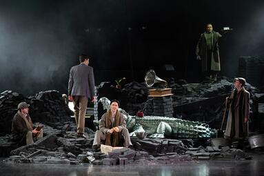 Vergrößerte Ansicht von Fünf Schauspieler stehen und sitzen auf einer Bühne, die wie eine zerstörte Landschaft aus dunklen Ziegeln gestaltet ist. In der Mitte liegt ein großes Krokodilmodell aus glänzendem Material. Ein Grammophon steht auf gestapelten Steinen. Hinten…