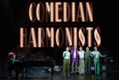 Vergrößerte Ansicht von Auf einer Theaterbühne stehen sechs Darsteller*innen vor einer großen, leuchtenden Beschriftung „COMEDIAN HARMONISTS“. Fünf von ihnen singen und posieren nebeneinander, teils mit Mikrofonen, während eine Person mit buntem Kopfschmuck links am Flügel…