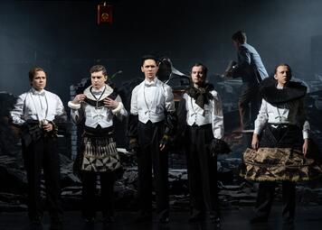 Vergrößerte Ansicht von Fünf Männer stehen nebeneinander auf einer Theaterbühne in Frack und weißem Hemd. Ihre Outfits sind durch ungewöhnliche Accessoires wie Röcke, Krägen oder Handschuhe auffällig verändert. Der Hintergrund zeigt eine zerstörte, ruinenartige Kulisse. Im…