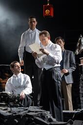 Vergrößerte Ansicht von In der Mitte steht ein Mann und weißer Veste, der von einem beschriebenen Blatt Papier liest. Um ihn herum stehen drei weitere, ähnlich gekleidete Männer und beobachten ihn. Sie wirken kritisch und angespannt, der Mann mit dem Papier unglücklich und…