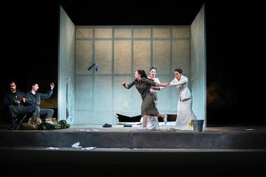 Vergrößerte Ansicht von Es befinden sich fünf Personen auf der Bühne, die aus zwei großen Stufen besteht. Auf dem Boden liegen Zettel verteilt und es stehen ein Metalleimer mit Gummihandschuhen, sowie Taschen und Stoffdecken rum. Im Hintergrund ist ein aus drei Wänden…