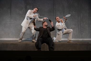 Vergrößerte Ansicht von Eine Person in braunem Anzug sitzt am Bühnenrand und hält zwei Glasflaschen in den Händen, die nach oben gerichtet sind. Den Kopf dreht die Person dabei zur Seite. Dahinter stehen zwei Personen in weißen Anzügen, sie halten beide einen…