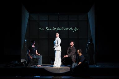 Vergrößerte Ansicht von Fünf Personen befinden sich auf der Bühne, eine im Hintergrund stehende ist dabei kaum zu erkennen, da sie vom Schatten verschluckt wird. Im Mittelpunkt steht eine Person in weißem Kostüm mit Rock und hochgesteckten Haaren, sie steht seitlich zum…