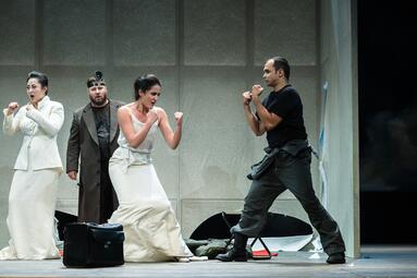 Vergrößerte Ansicht von Es sind vier Personen zu sehen, zwei davon tragen einen weißen Rock, einer ist in Arbeitskleidung und einer im braunen Anzug. Drei Personen ballen die Hände zu Fäusten und haben sie in Abwehrhaltung vor ihrem Körper platziert. Zwei der Personen…