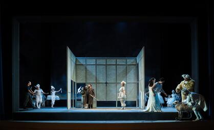 Vergrößerte Ansicht von Es befinden sich viele Personen auf der Bühne, gekleidet in Kleidern und Anzügen. Viele haben weiße hochtoupierte Perücken auf. Dazwischen sind wieder die Personen im weißen Rock, Arbeitskleidung und im braunen Anzug zu sehen.