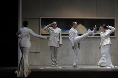 Vergrößerte Ansicht von Vier Personen in weißen Anzügen stehen in ungewöhnlichen Körperhaltungen vor einem langen Gemälde. Eine Person kehrt der Kamera den Rücken zu und hat die Arme in Tanzhaltung gehoben, die Person daneben salutiert, rechts davon hebt einer sein Bein…