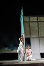 Vergrößerte Ansicht von Zwei Personen in weißen Kleidern aus Tüll sind im Mittelpunkt des Bildes, getrennt durch eine Wand. Eine Person räkelt sich an dieser Wand, die Person auf der anderen Seite kniet und singt.