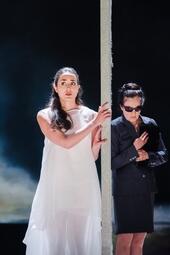 Vergrößerte Ansicht von Zwei Personen getrennt durch eine Wand. Eine Person in weißem Tüllkleid, eine in schwarzem Anzug mit Rock und dunkler Sonnenbrille. Im Hintergrund sind Wolken oder Nebel zu sehen.