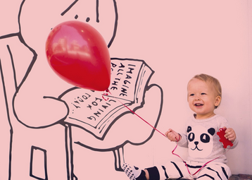 Vergrößerte Ansicht von Ein Kleinkind sitzt lachend vor einer Wand mit Comic-Motiv und hält einen roten Luftballon an einer Schnur.