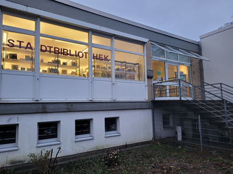 Vergrößerte Ansicht von Von außen: Die hell erleuchtete Stadtteil-Bibliothek Neckarau in der Dämmerung.