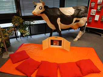 Vergrößerte Ansicht von Ein Foto zeigt ein Kamishibai-Theater auf dem Boden. Davor liegen sechs Kissen im Halbkreis, dahinter steht ein lebensgroßes Modell der Bilderbuchfigur &quot;Kuh Lieselotte&quot;.