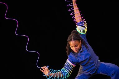 Vergrößerte Ansicht von Es ist eine Frau zu sehen in einem einem dunkelblauen Hemd, die große bunte Spiralen um ihre Arme trägt. Die Spiralen sind länger als die Arme der Frau.